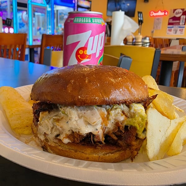 A pulled pork sandwich with creamy sauce and chips on a plate, with a pink 7UP can in the background.