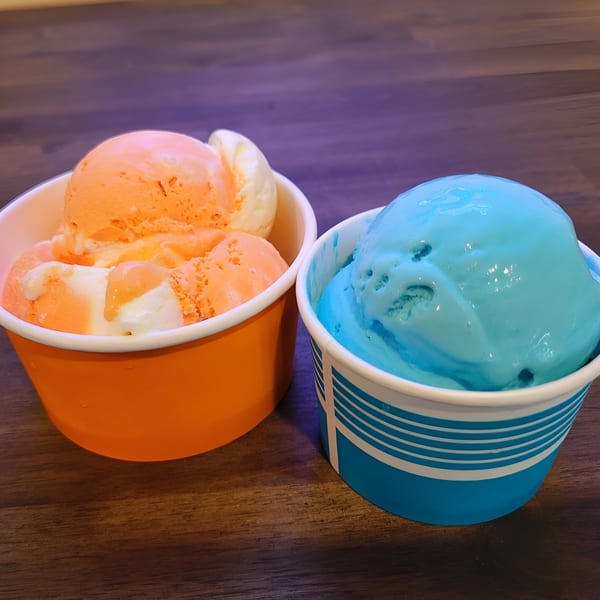 Two cups of ice cream, one orange and white, the other blue, on a wooden table.